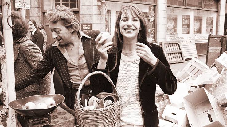 Jane Birkin walking through a Parisian street in the 1970s holding a straw basket